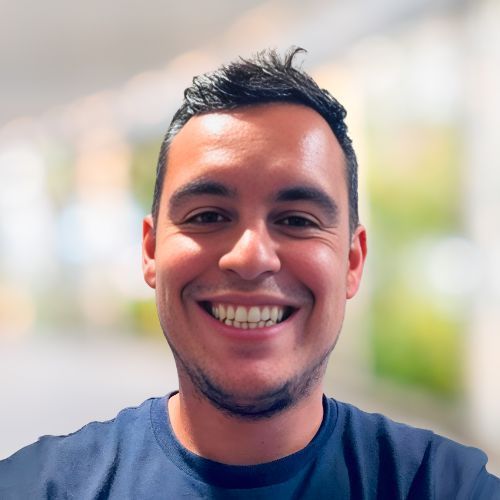 Man with dark hair smiling, wearing a blue shirt. Blurred, bright background.
