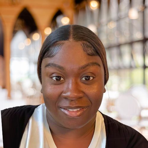 Woman with dark hair smiles, wearing a white top and black cardigan, in a cafe.