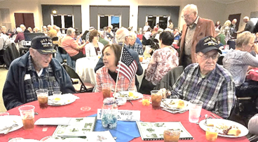 A group of people are sitting at tables with plates of food and drinks celebrating Veterans Day