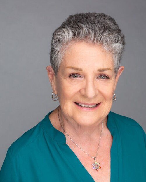 An older woman wearing a blue shirt and a necklace is smiling for the camera.