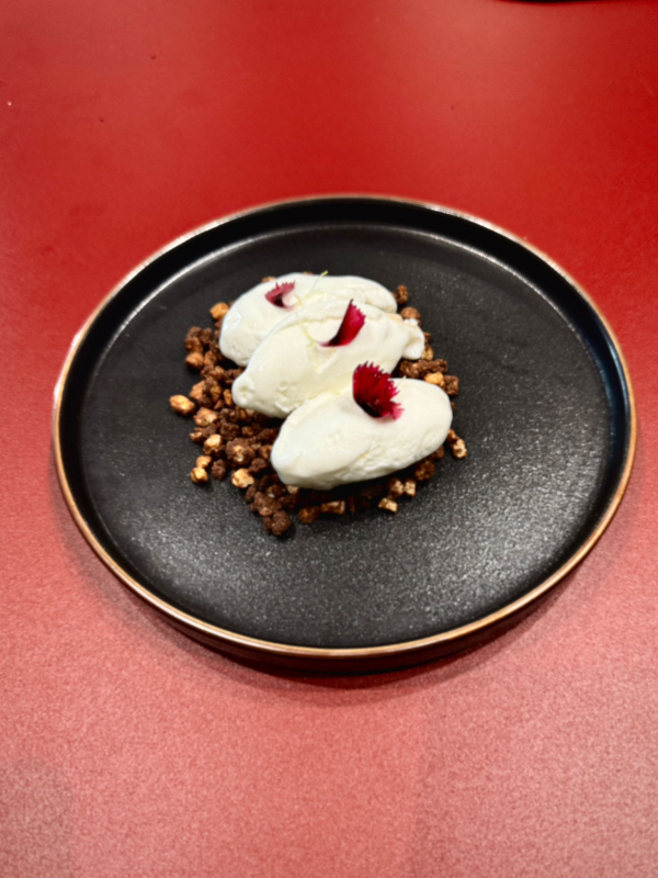 Dessert: tre palline bianche su croccante di cioccolato, guarnite di rosso su un piatto nero con bordi dorati.