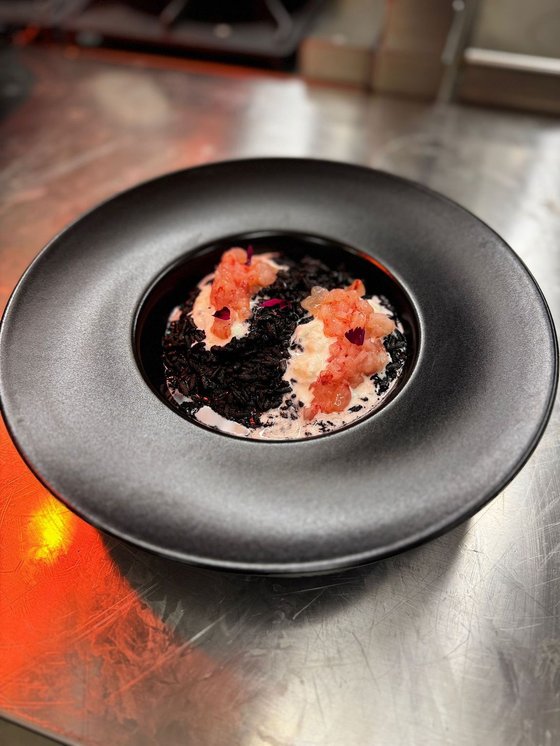 Piatto nero con riso nero e cibo dalle tonalità rosa, forse salmone, e petali di fiori.