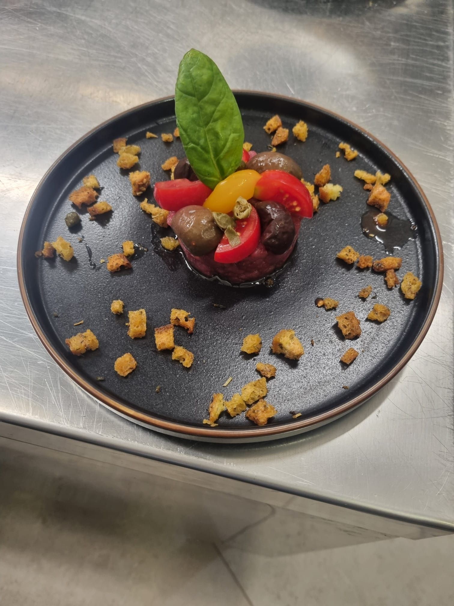 Piccolo piatto scuro con antipasto di barbabietola e pomodoro, circondato da crostini. Sopra, una foglia di spinaci.