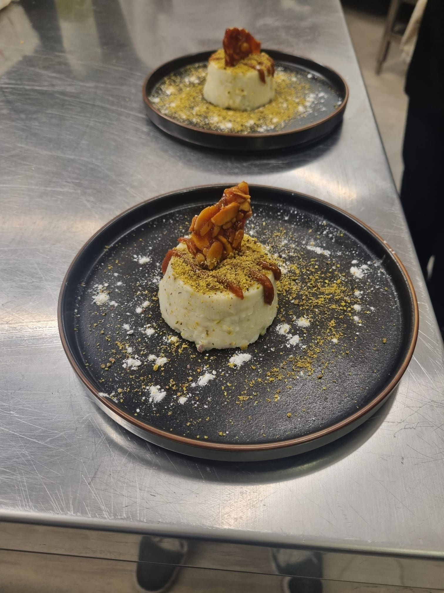 Due dessert su piatti neri, con montature bianche, guarniti con noci e salsa, su una superficie argentata.