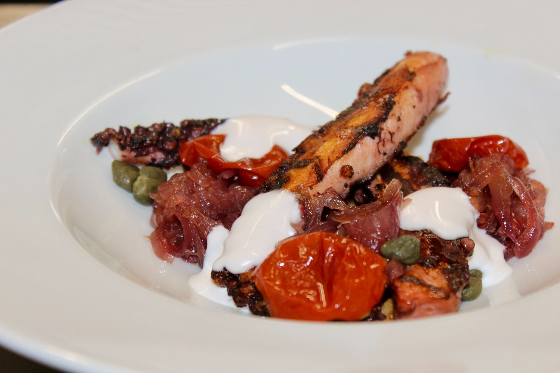 Polpo alla griglia con pomodori, cipolla rossa, capperi e salsa bianca in una ciotola bianca.