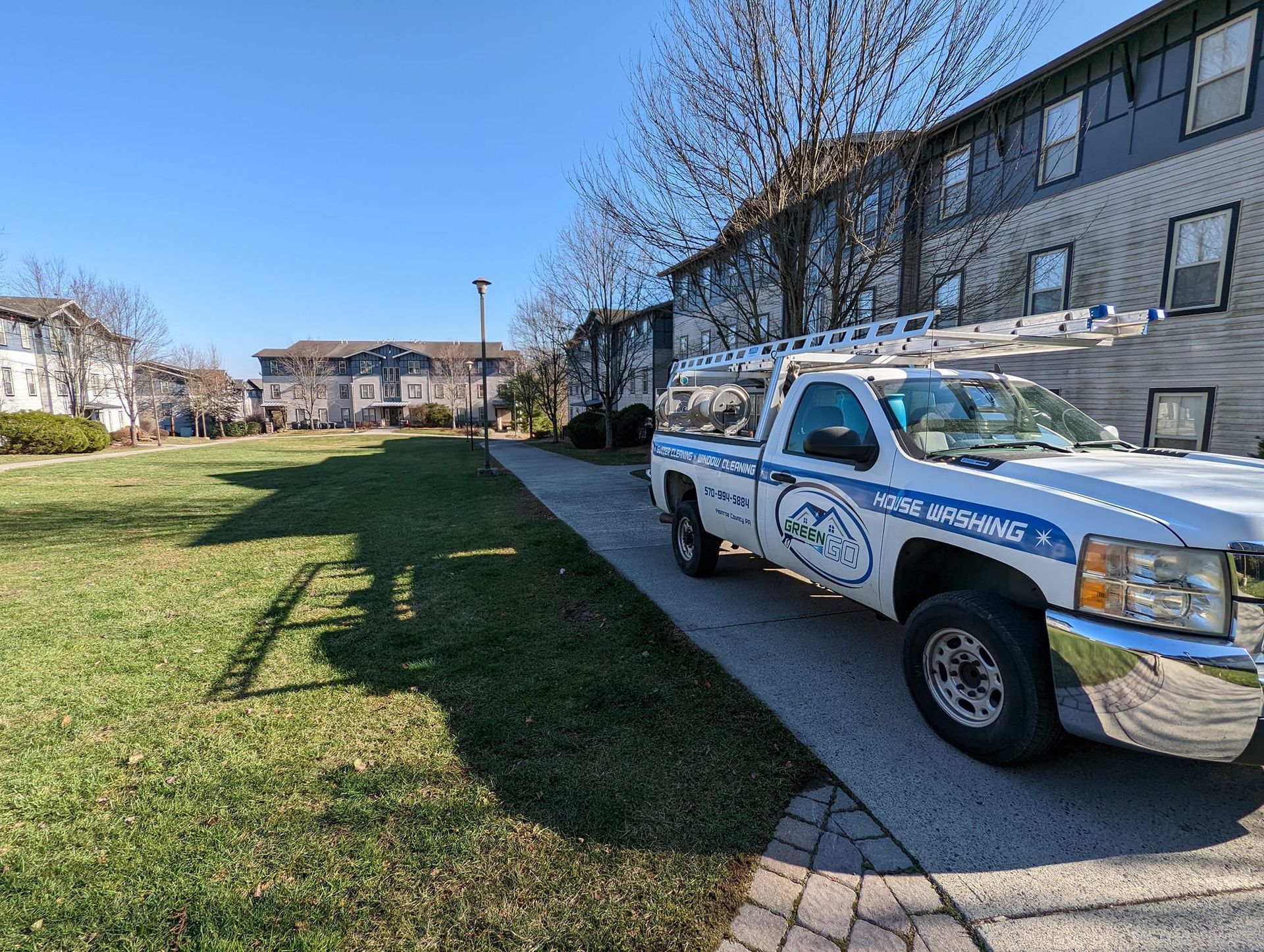White work truck parked on a sidewalk beside grass, in front of an apartment building on a sunny day.