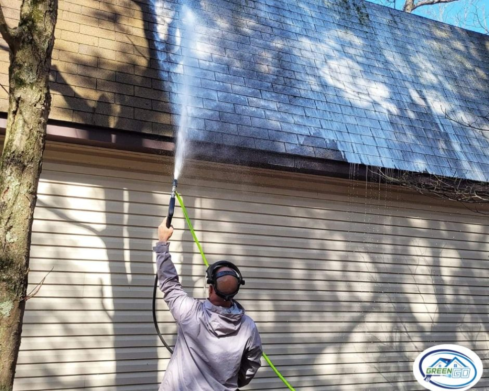 Person pressure washing a roof. The setting is outdoors with a tree and beige siding.