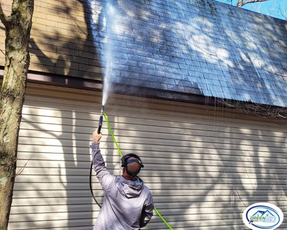 Person pressure washing a roof. The setting is outdoors with a tree and beige siding.