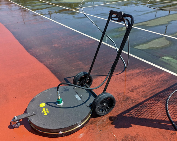 Pressure washer cleaning a red tennis court. Black machine with wheels, partially cleaned surface.