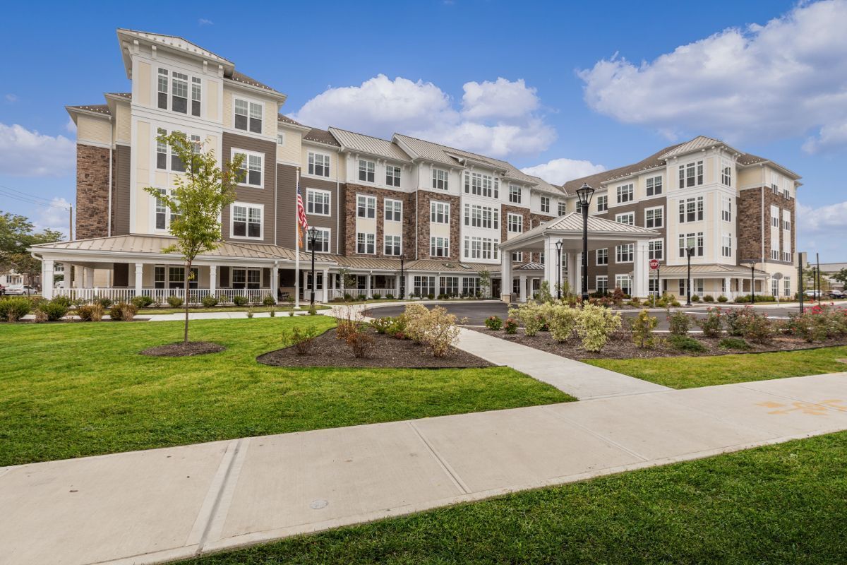 Multi-story senior living facility with tan and brick facade, green lawn, blue sky.