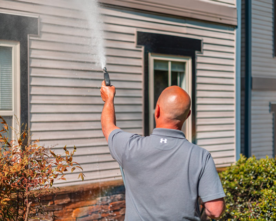 Man power washing a house with gray siding.