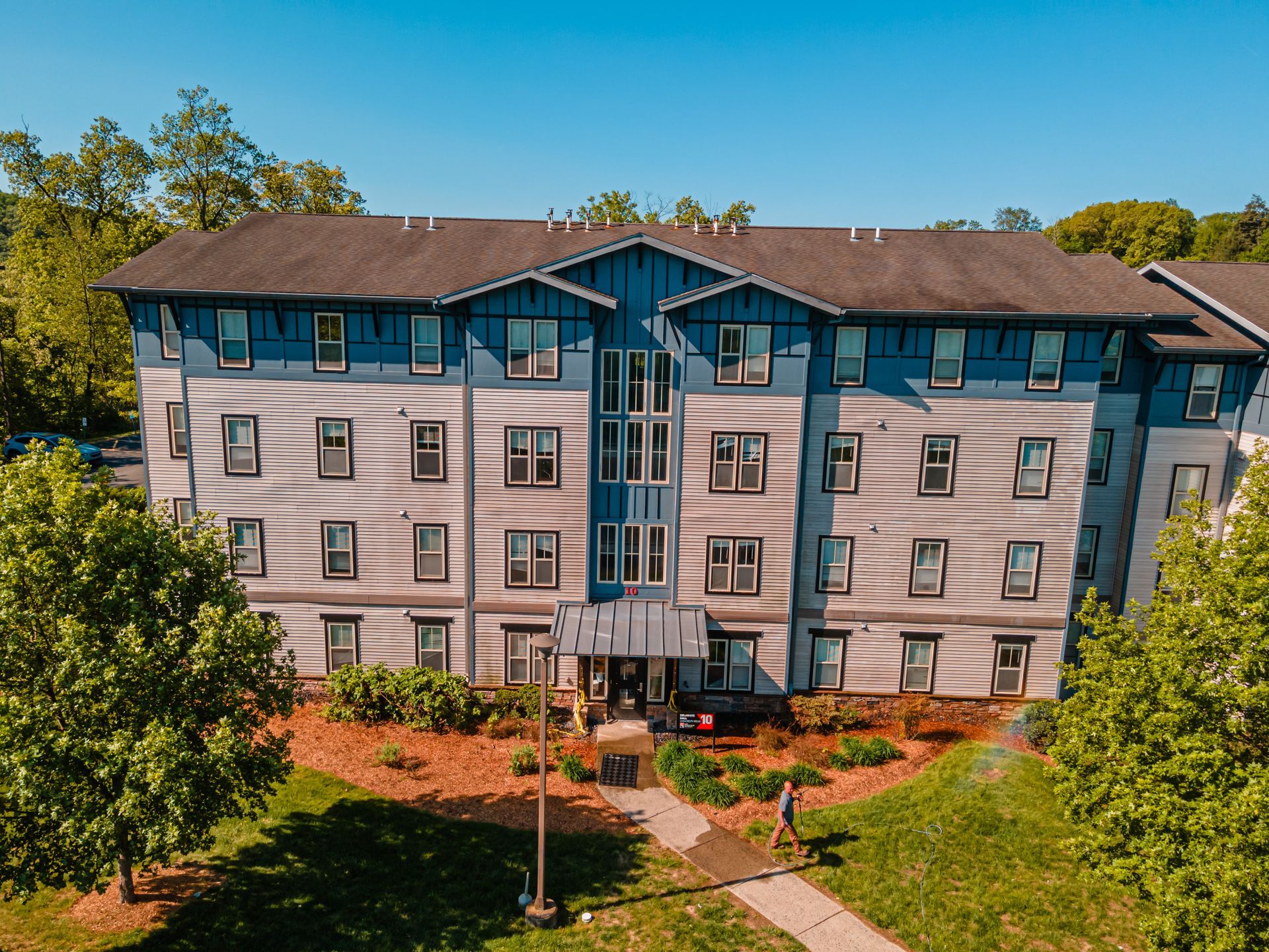 Multi-story apartment building with blue and gray siding, surrounded by grass and trees, sunny day.