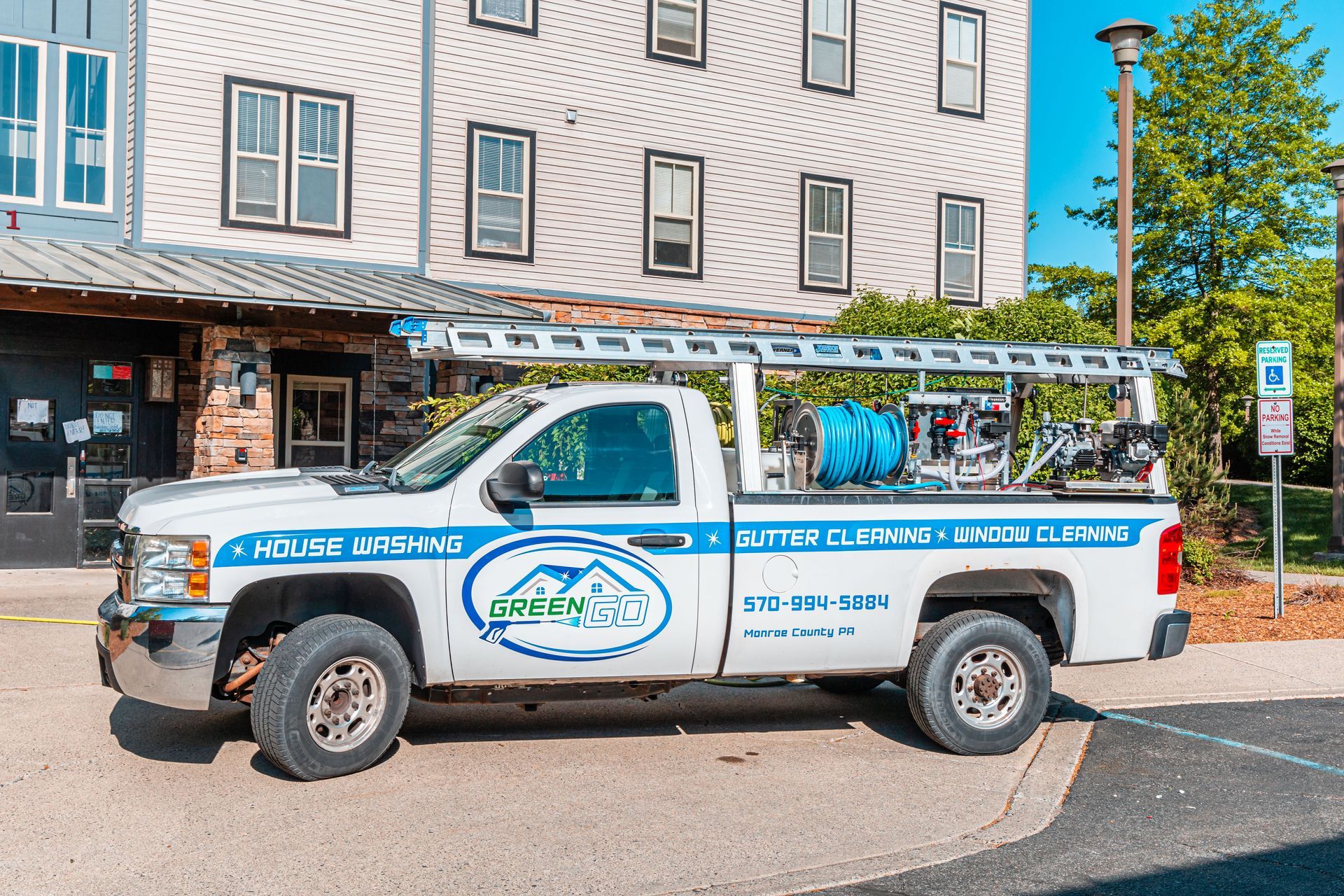 White truck with a ladder on top, parked in front of a building. The truck has a company logo on the side, 