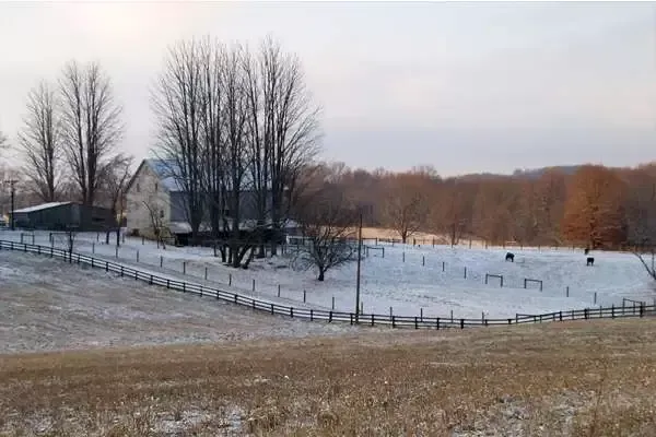 Snow-covered rural landscape with a barn and a fence. Bare trees and a field in the foreground, woods in the background.