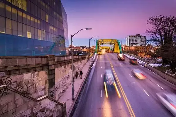 City street at dusk, blurred car headlights on roadway under a green bridge, pedestrians on sidewalk, purple sky.