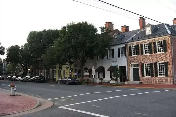 Street with brick buildings, trees, and parked cars under a partly cloudy sky.