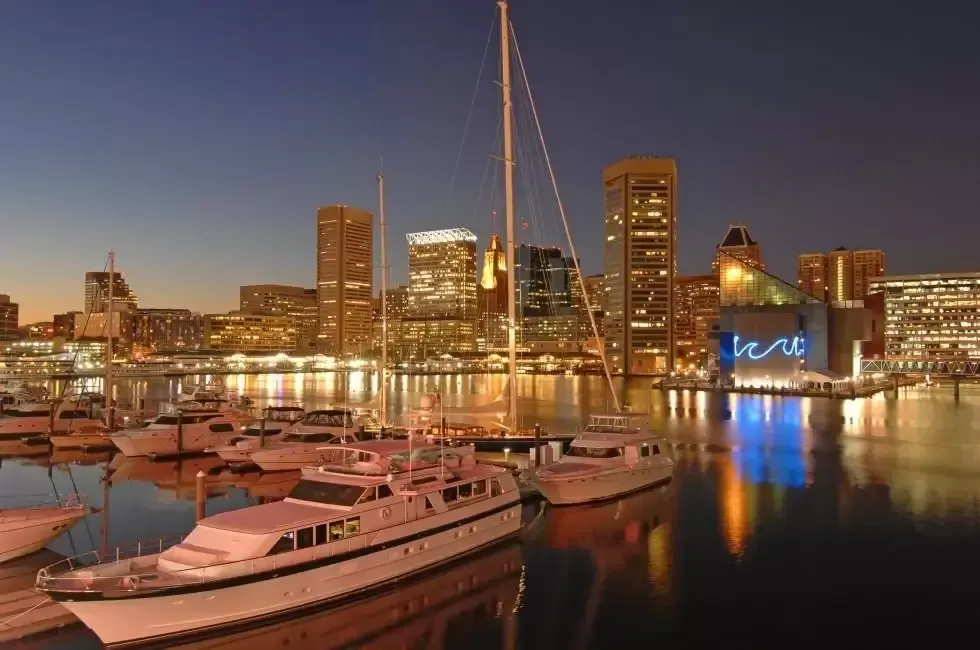 Nighttime view of Baltimore's Inner Harbor with lit skyscrapers reflecting in the water, boats in foreground.