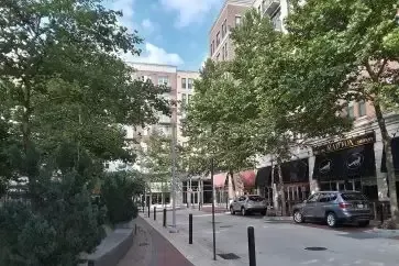 Street view of apartment building and shops, lined with trees and cars parked on the side.