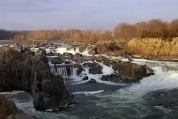 Rushing river cascades over rocky ledges. Sunlight highlights water and surrounding forest.