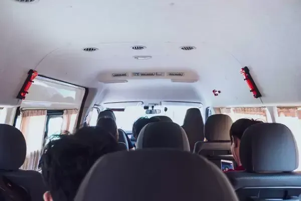 Interior of a van with passengers. Gray seats, white ceiling, and some windows are visible.