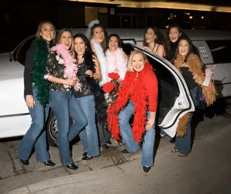 Women in boas and jeans pose in front of a white limousine at night; smiling and laughing.