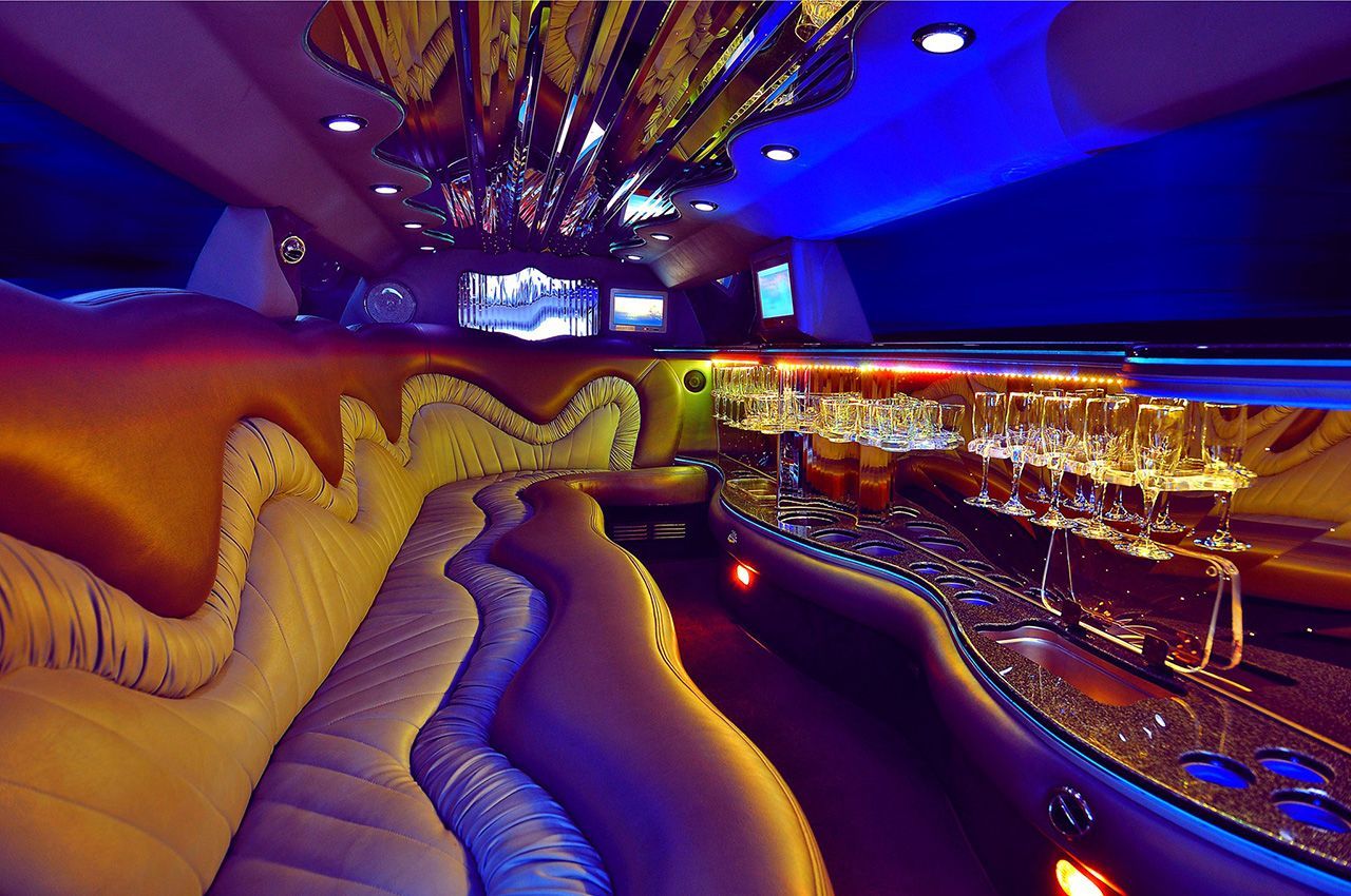 Interior of a limousine with curved seating, a lit bar, and blue and gold lighting.