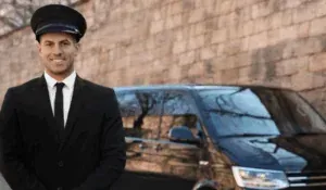 Man in chauffeur uniform stands near a black van, smiling. Background: brick wall.