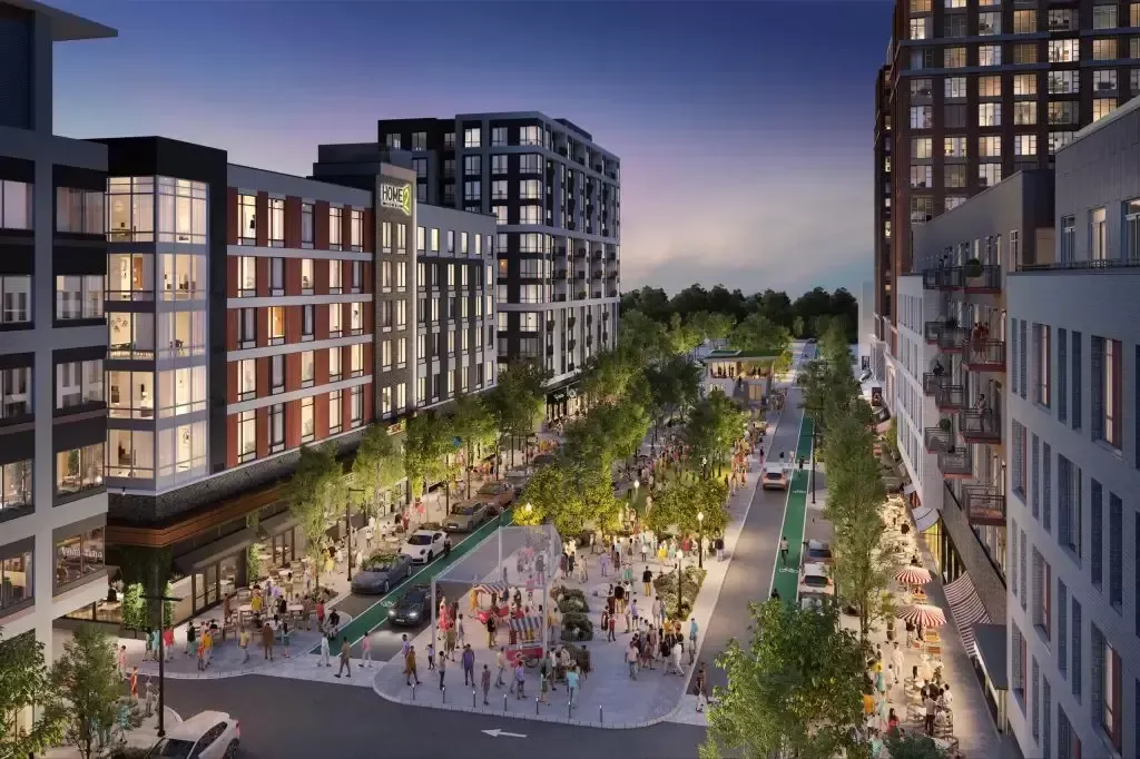 Nighttime view of a city street with pedestrians, buildings, trees, and a central park-like area with lights.