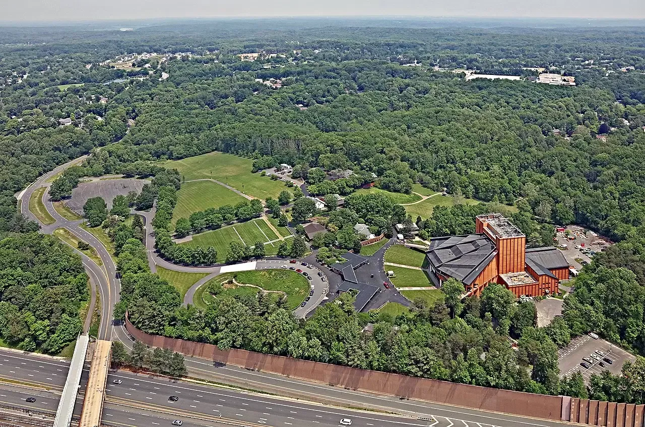 Aerial view of a large, brown building nestled among trees and a green field; road and highway in foreground.