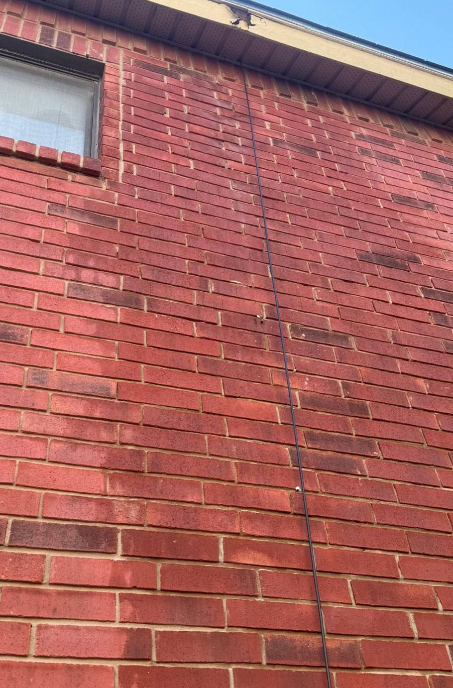 A long, dark wire runs vertically down the center of a red brick wall, next to a window on the upper left.