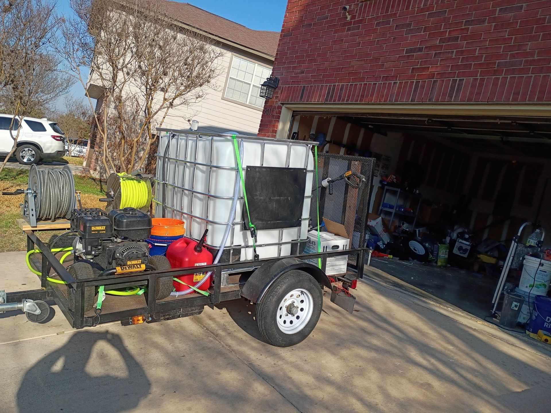 A utility trailer carrying a large water tank, a power washer, and fuel cans sits on a driveway in front of a garage.