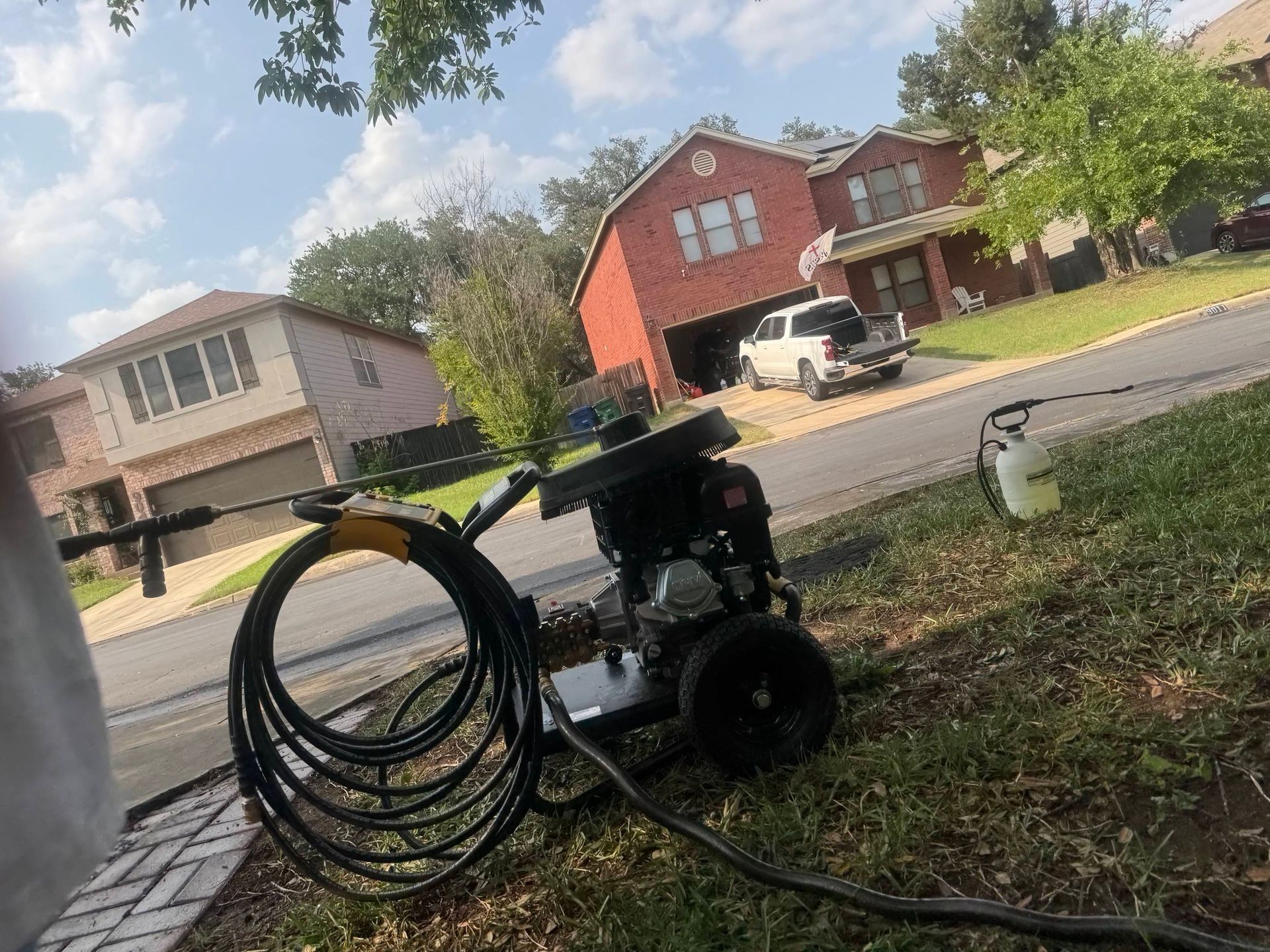 A pressure washer with a coiled black hose sitting on grass near a residential street with houses in the background.
