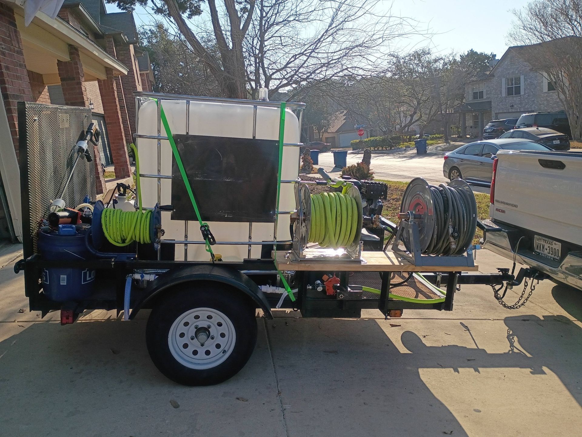 A utility trailer with a large water tank, pressure washer equipment, and two hose reels, attached to a pickup truck.