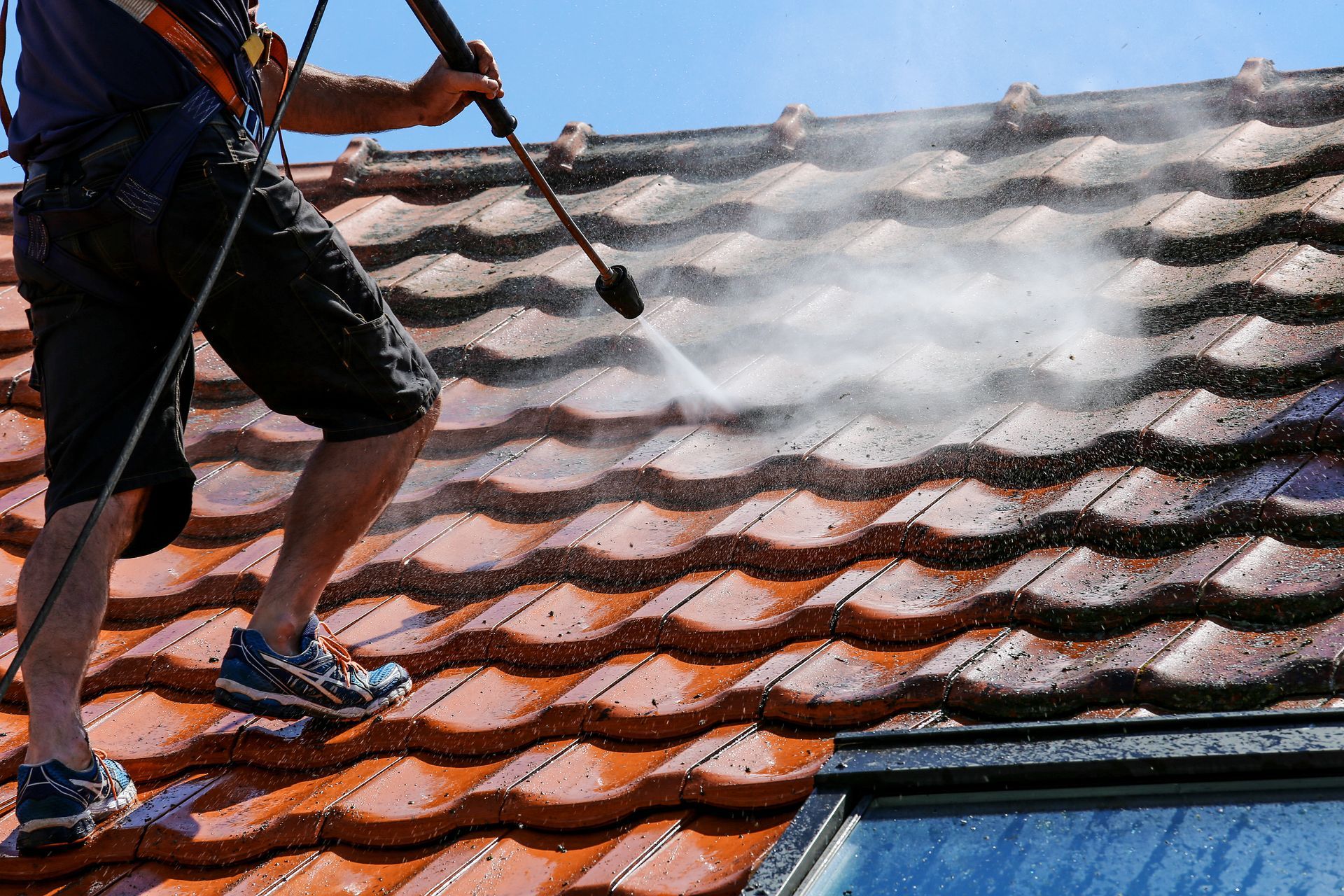 A person uses a pressure washer to clean dirty brown roof tiles under a clear blue sky.