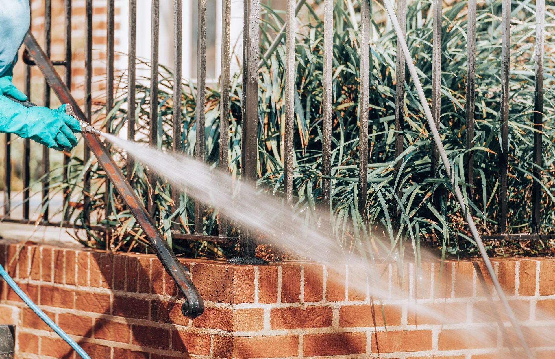 A person wearing green gloves power washes a brick wall and metal fence.