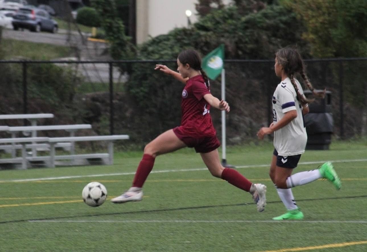 Two SCOR girls are playing soccer on a field with a green flag in the background
