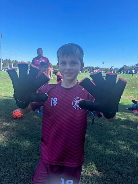 A young SCOR boy wearing a maroon shirt and black gloves is standing on a soccer field.