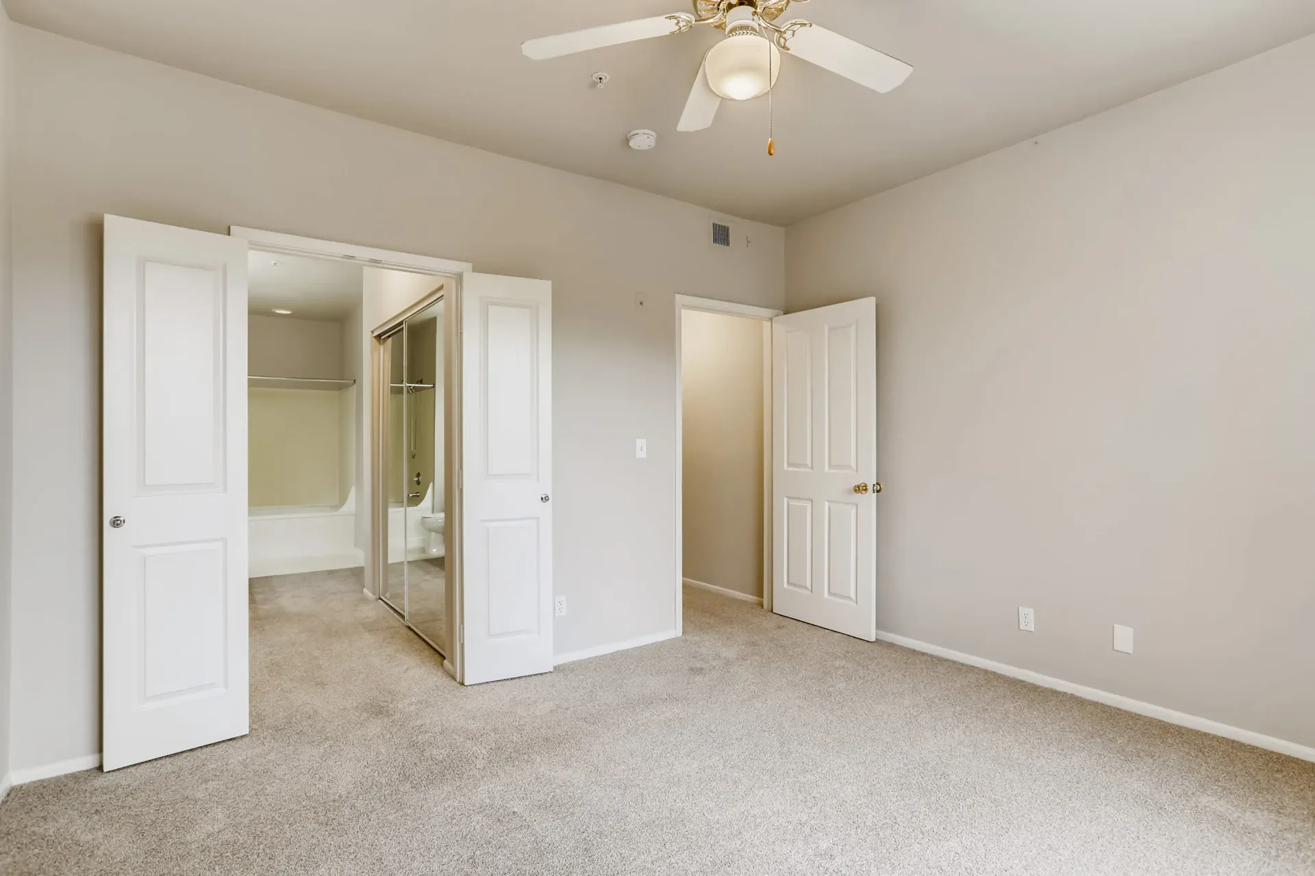 Bedroom with beige walls, white doors, ceiling fan, and beige carpet; closet and bathroom door visible.