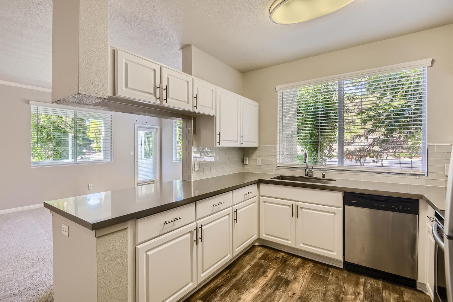Bright kitchen with white cabinets, gray countertops, stainless steel dishwasher, and a large window over the sink.