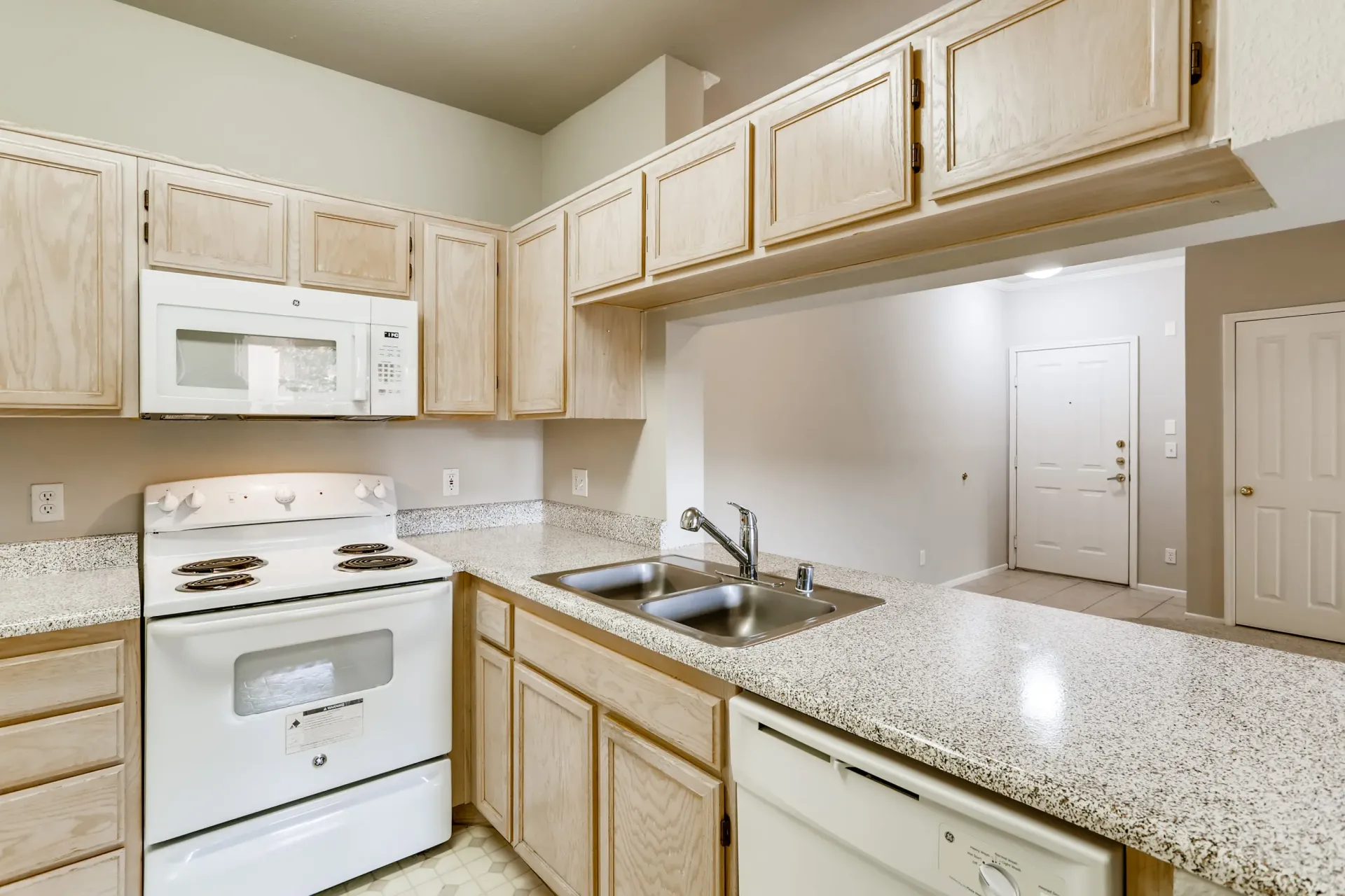 Apartment kitchen with light wood cabinets, white stove and microwave, double sink, and speckled countertops.