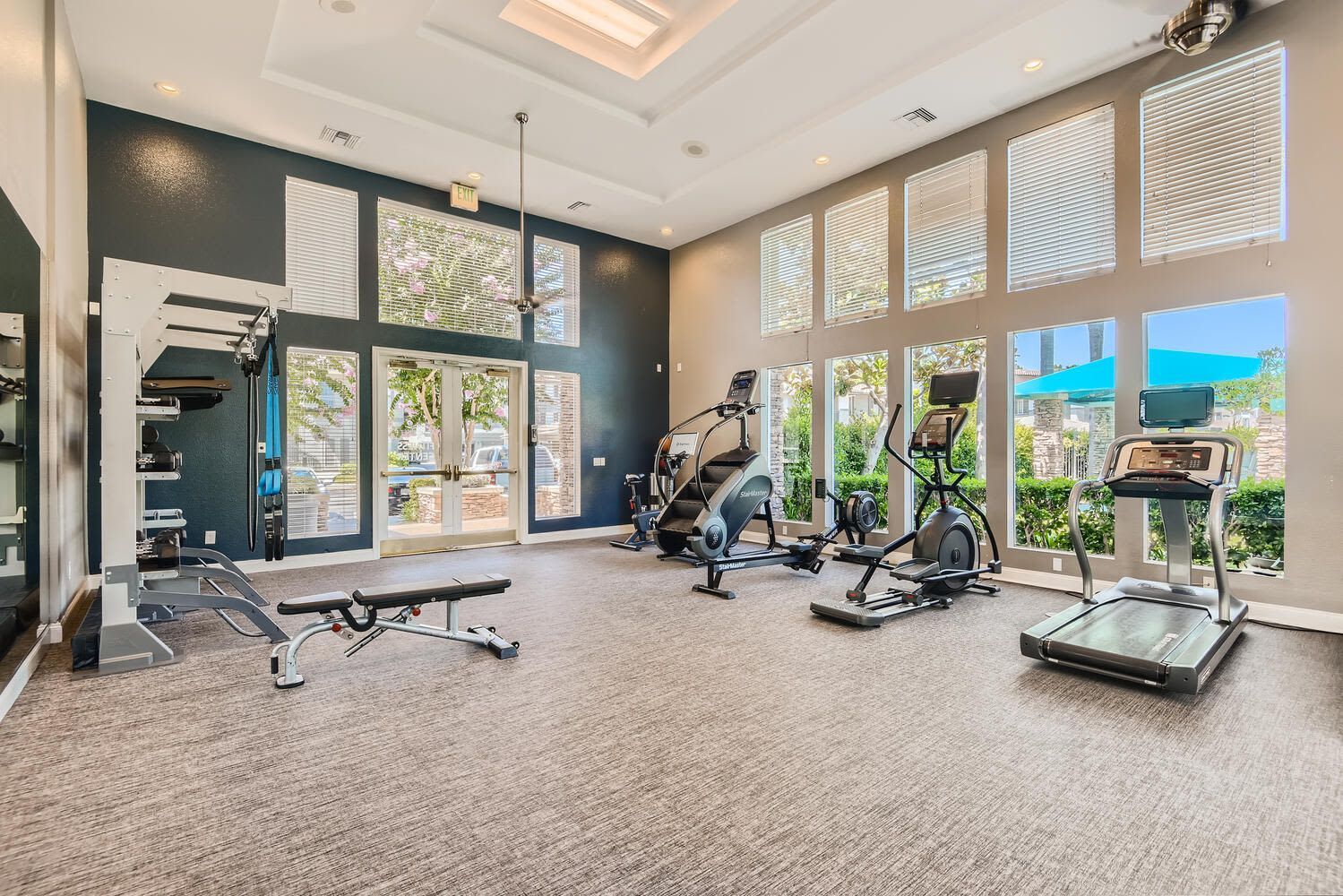 Bright apartment community gym with cardio machines and a weight station by large windows.