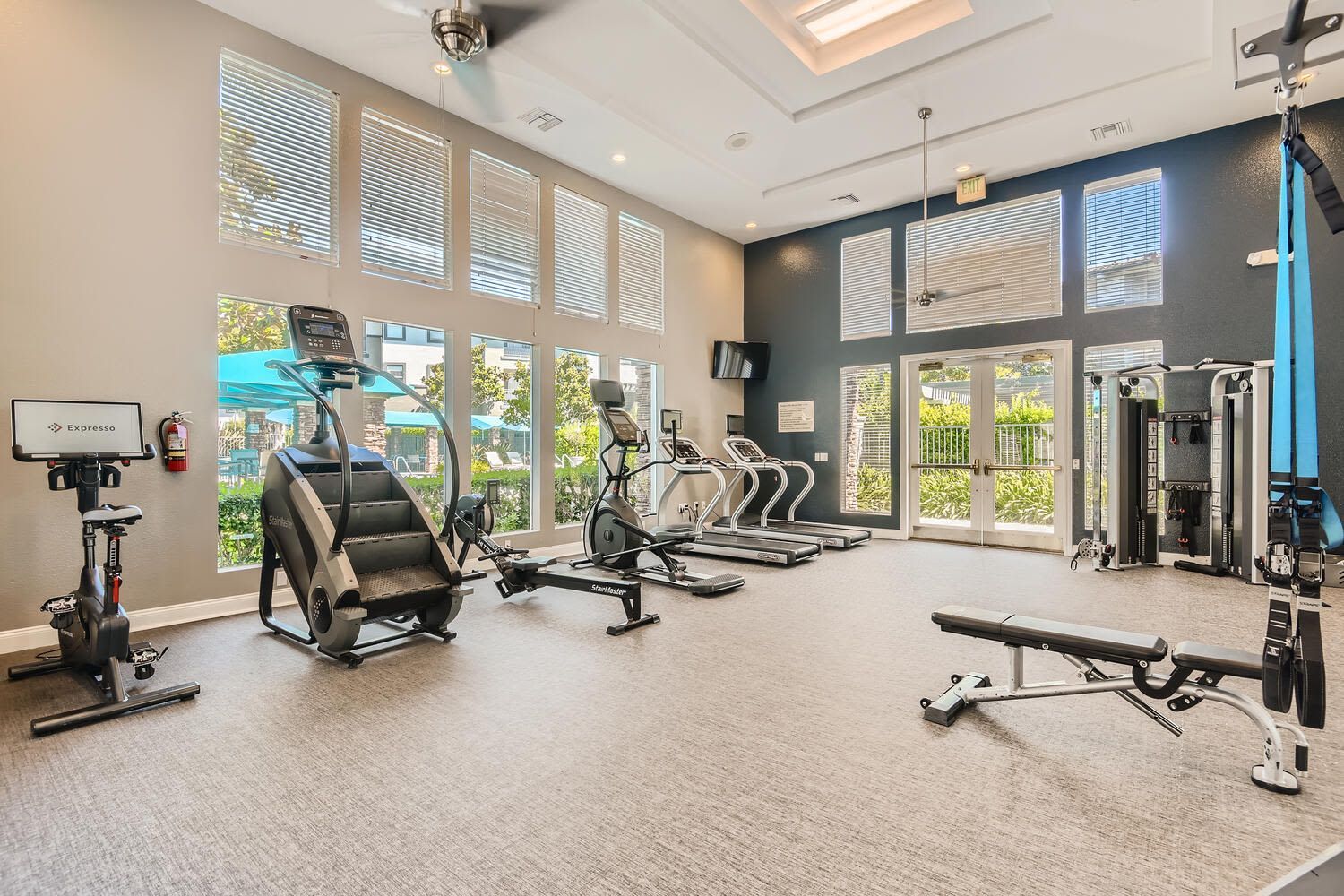 Bright apartment fitness center with treadmills, bikes, and weight machines beside large windows.