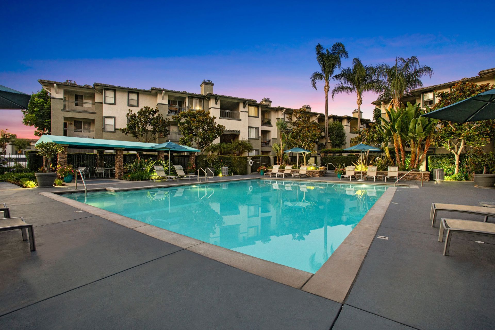 Outdoor apartment community pool with lounge chairs, umbrellas, and surrounding buildings.