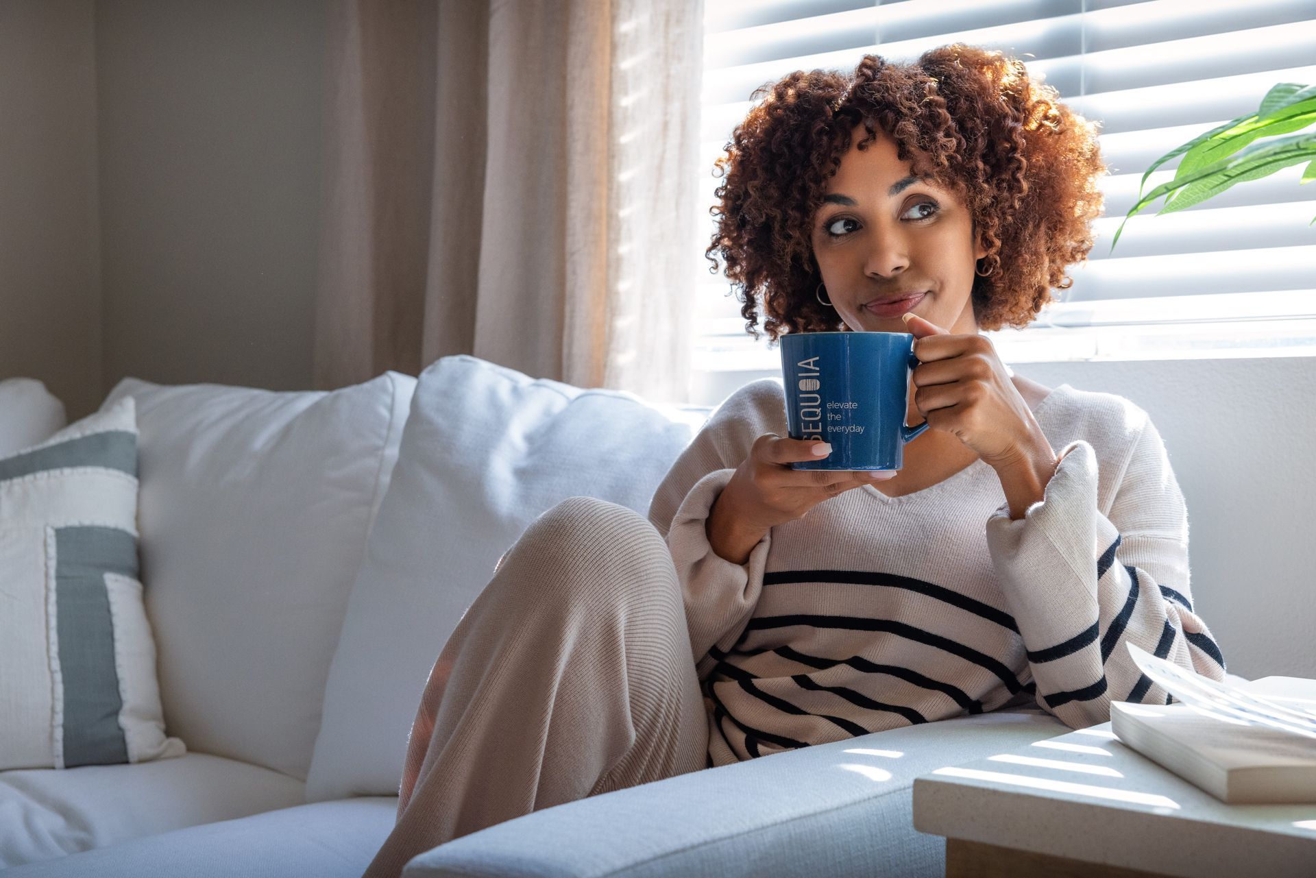 Woman sitting on a couch, holding a mug, looking out the window, sunlight.