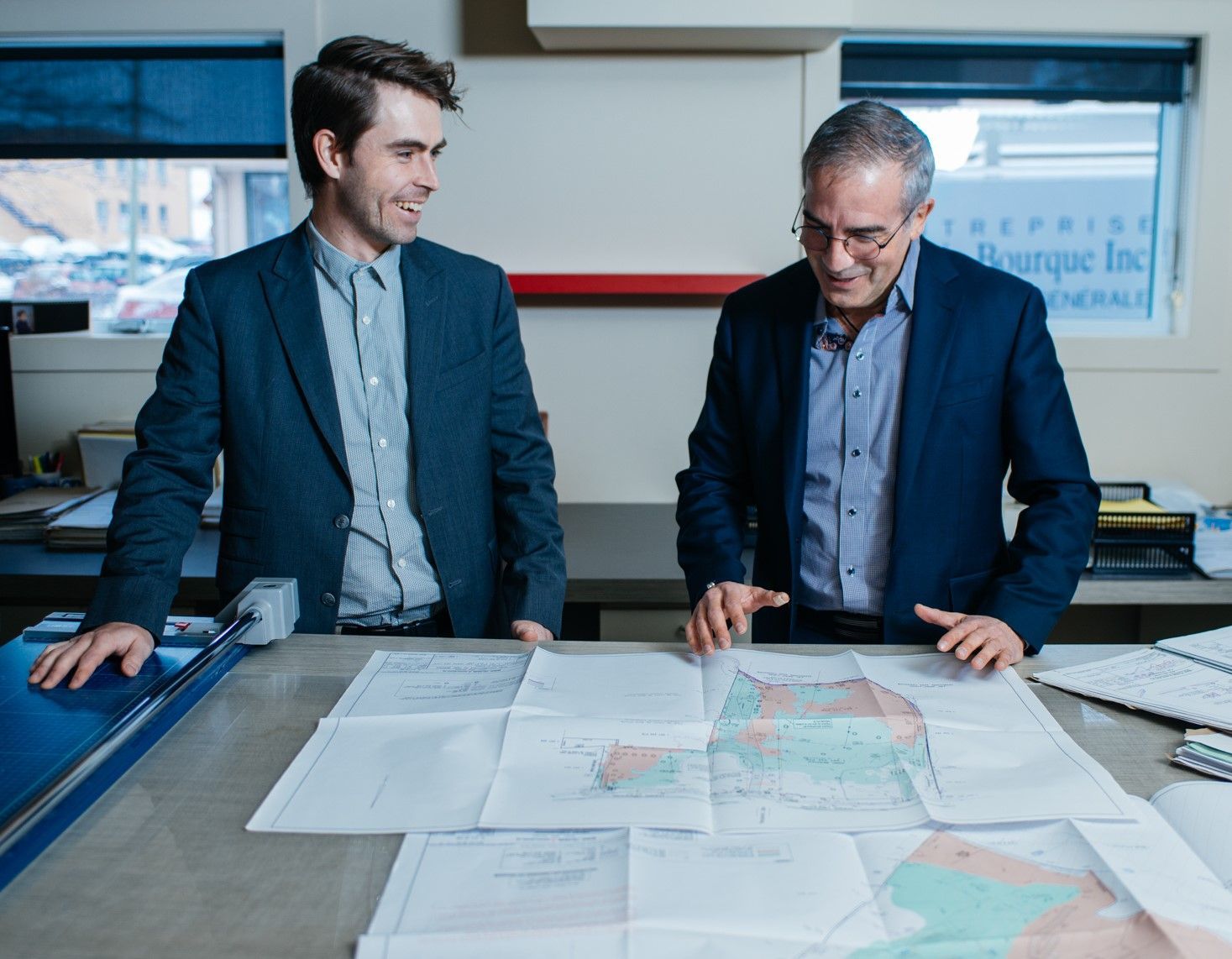 Deux hommes examinent des plans à un bureau. L'un sourit, l'autre montre du doigt.