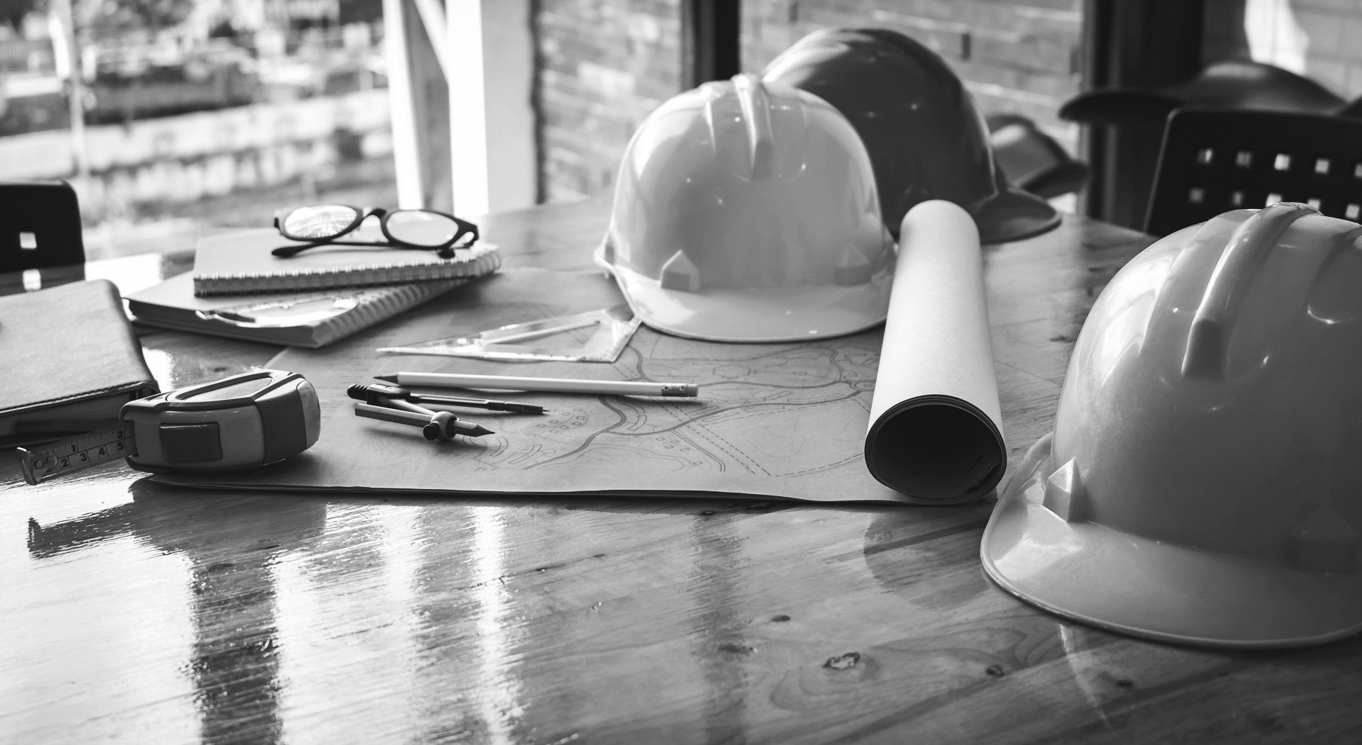 A black and white photo of hard hats , a blueprint , and other construction tools on a wooden table.