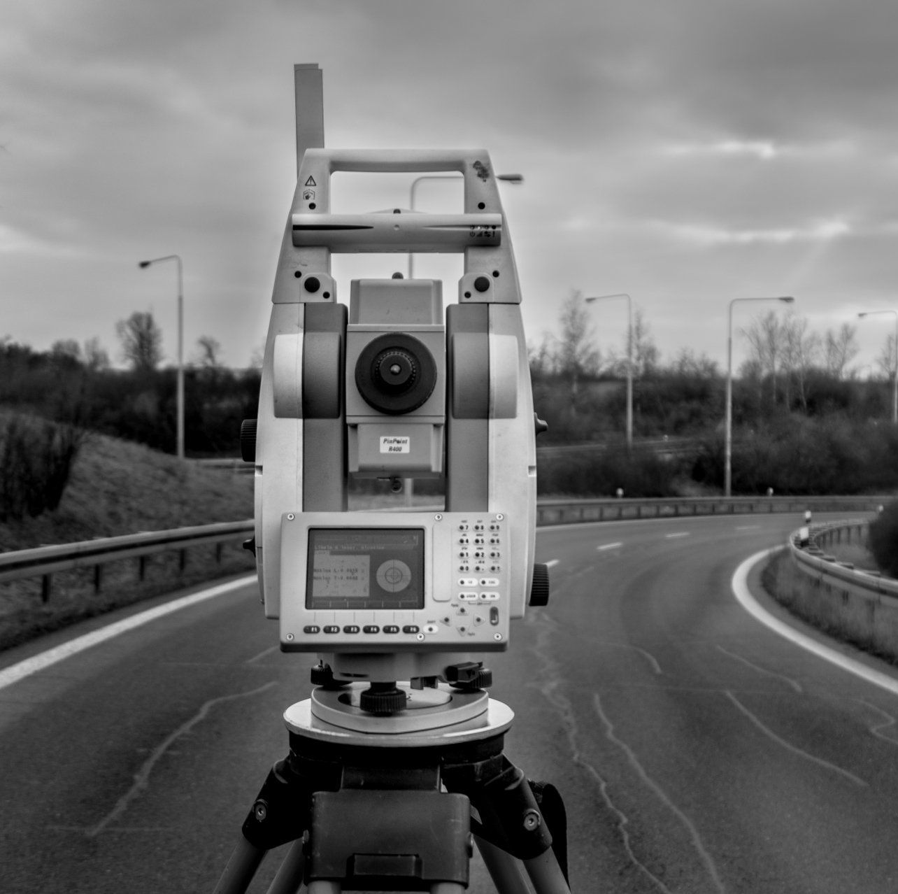 Station totale de levé topographique sur trépied, positionnée sur une route avec une voie courbe et des arbres en arrière-plan.