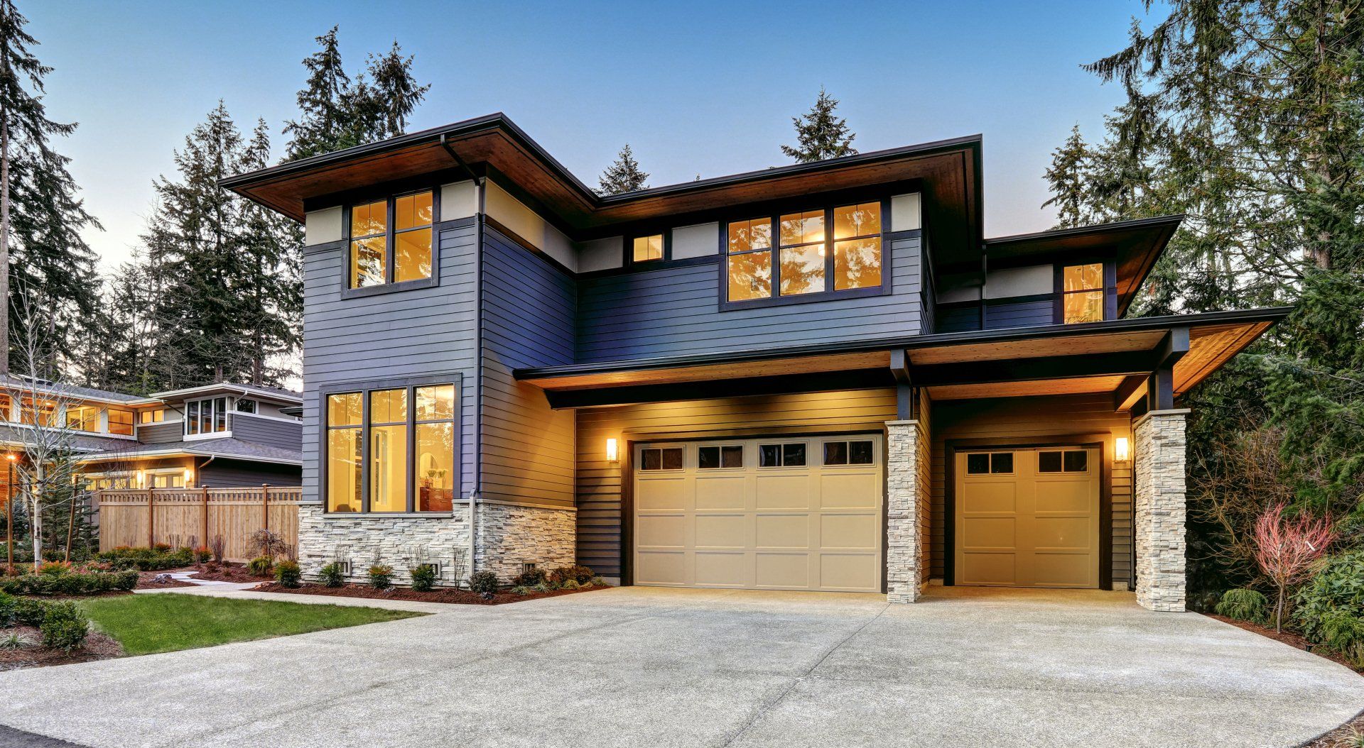 Maison moderne à deux étages avec revêtement bleu, accents de pierre et garage pour deux voitures, située dans une zone boisée.
