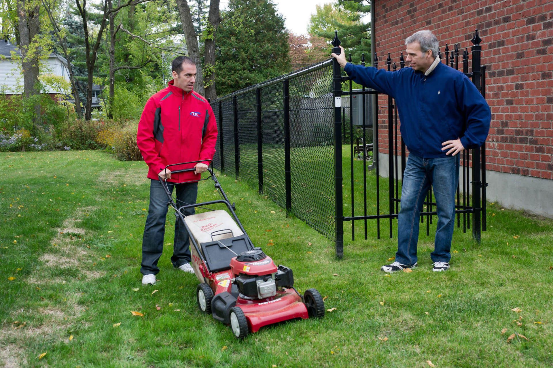 Un homme tond la pelouse tandis qu'un autre s'appuie contre une clôture noire à côté d'un bâtiment en briques.