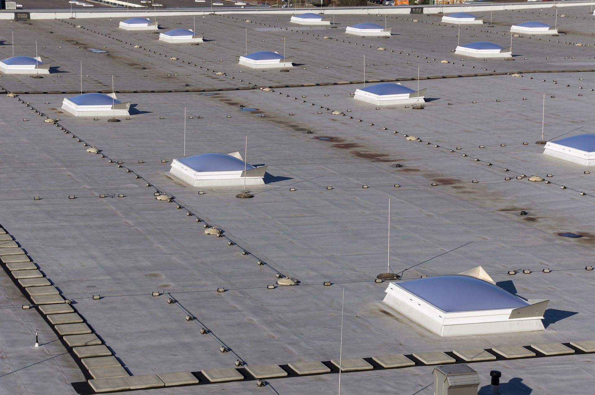 Eine Luftaufnahme eines flachen Gewerbedaches mit mehreren weißen Oberlichtern und Blitzableitern, die in Reihen angeordnet sind und sich von einer dunklen Oberfläche abheben.